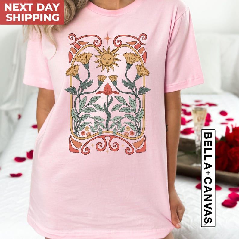 May include: Pink t-shirt with a vintage-style floral design featuring a sun face, yellow flowers, and red flowers. The design is surrounded by a decorative frame.