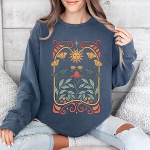 Op de afbeelding: Een marineblauw sweatshirt met een vintage-ge&iuml;nspireerde afbeelding van een zon en bloemen in een bloemvormige boog. De afbeelding is in tinten geel, oranje, rood en groen.