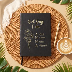 May include: A black journal with gold text reading God Says I Am and the name ANNA with corresponding words Alive, Noble, New, and Adored, accompanied by a gold pen and a floral line art illustration on the front cover.