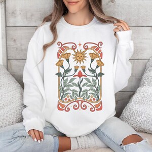 Op de afbeelding: Witte trui met een grafische print van een gestileerde zon en bloemen in een vintage art nouveau-stijl.