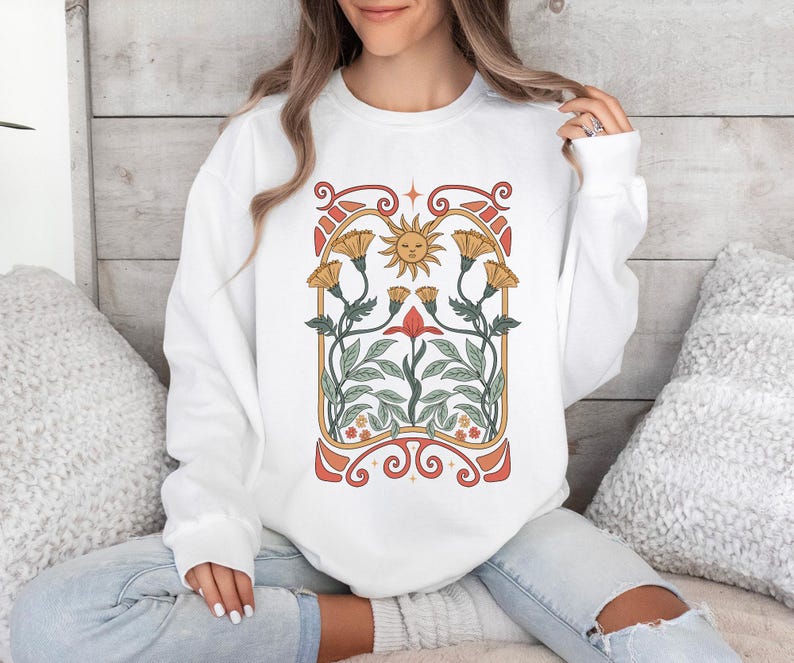 Op de afbeelding: Witte trui met een grafische print van een gestileerde zon en bloemen in een vintage art nouveau-stijl.