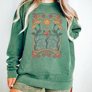 Op de afbeelding: Een groen sweatshirt met een vintage-stijl grafische afbeelding van een zon en bloemen. De afbeelding is in tinten oranje, geel, rood en groen.