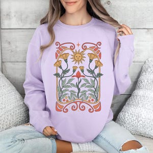 Op de afbeelding: Een lavendelkleurig sweatshirt met een grafische print van een zon en bloemen in een vintage art nouveau-stijl.