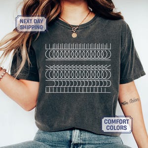 May include: Dark gray t-shirt with a white geometric design. The design features a series of horizontal lines with various patterns, including loops and zigzags. The shirt has the word "PEPPER" printed at the bottom.