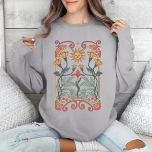 Op de afbeelding: Een grijs sweatshirt met een bloemenontwerp met een zon, bloemen en bladeren. Het ontwerp is in vintage art nouveau-stijl.