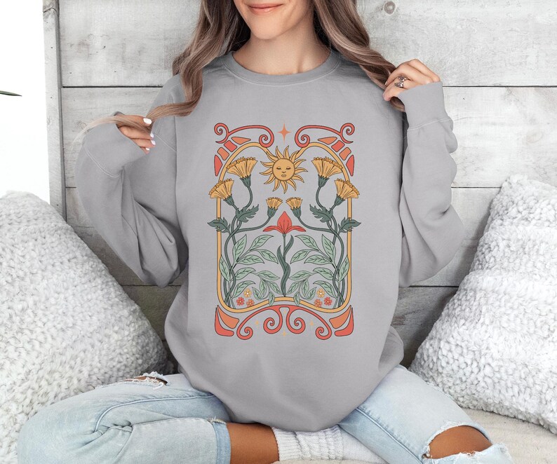 Op de afbeelding: Een grijs sweatshirt met een bloemenontwerp met een zon, bloemen en bladeren. Het ontwerp is in vintage art nouveau-stijl.