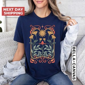 May include: A navy blue t-shirt with a vintage-style graphic of a sun and flowers in a decorative frame. The graphic features a yellow sun with a smiling face, orange and yellow flowers, and green leaves. The frame is made of a dark red, orange, and yellow design.