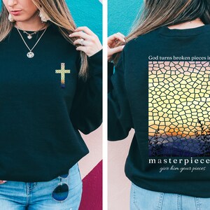 May include: Black sweatshirt with a gold cross on the front and a stained glass sunset design on the back. The back design includes the text "God turns broken pieces into masterpieces give him your pieces".