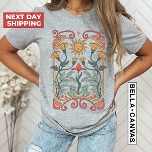 May include: Gray t-shirt with a graphic of a sun, flowers, and leaves in an art nouveau style. The graphic is in shades of orange, yellow, red, and green.