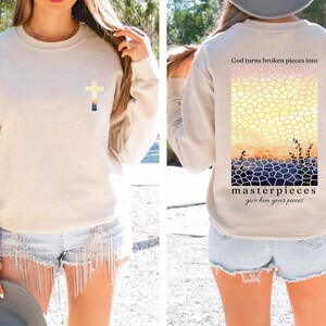 May include: A beige sweatshirt with a cross on the front and a mosaic sunset design on the back. The back design includes the text "God turns broken pieces into masterpieces give him your pieces".