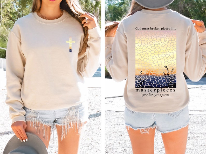 May include: A beige sweatshirt with a cross on the front and a mosaic sunset design on the back. The back design includes the text "God turns broken pieces into masterpieces give him your pieces".