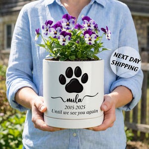 May include: A white cylindrical ceramic planter containing purple and white pansies. The front features a black paw print graphic above the name "mila", the years "2015-2025", and the text "Until we see you again" in a black script font.