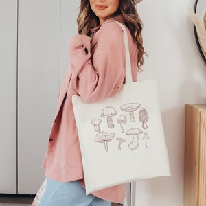 May include: A white tote bag with a brown line drawing of various mushrooms. The bag has a long strap and is being held by a person wearing a pink sweater and blue jeans.