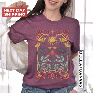 May include: A purple t-shirt with a graphic design of a sun, flowers, and leaves in a vintage art nouveau style.