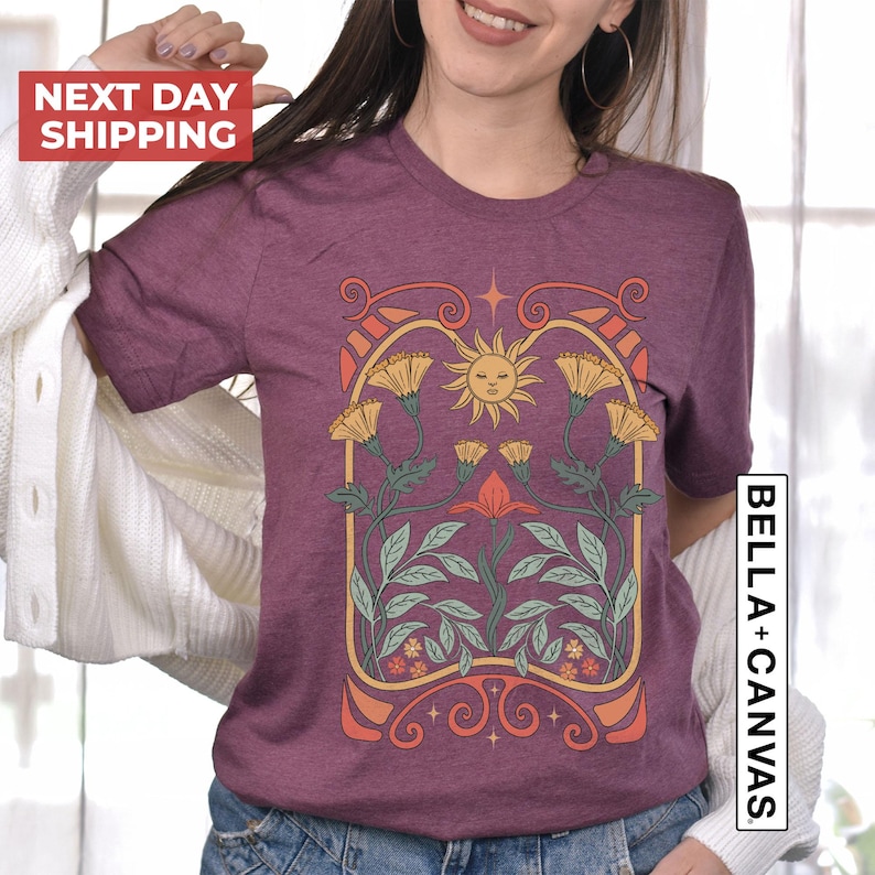 May include: A purple t-shirt with a graphic design of a sun, flowers, and leaves in a vintage art nouveau style.