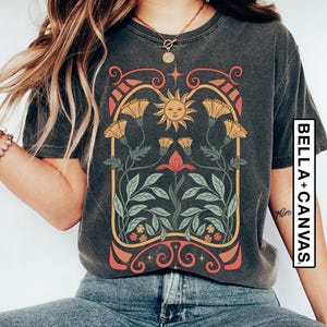 May include: A dark grey t-shirt with a vintage-style graphic design featuring a sun with a face, flowers, and leaves. The design is framed by a decorative border.