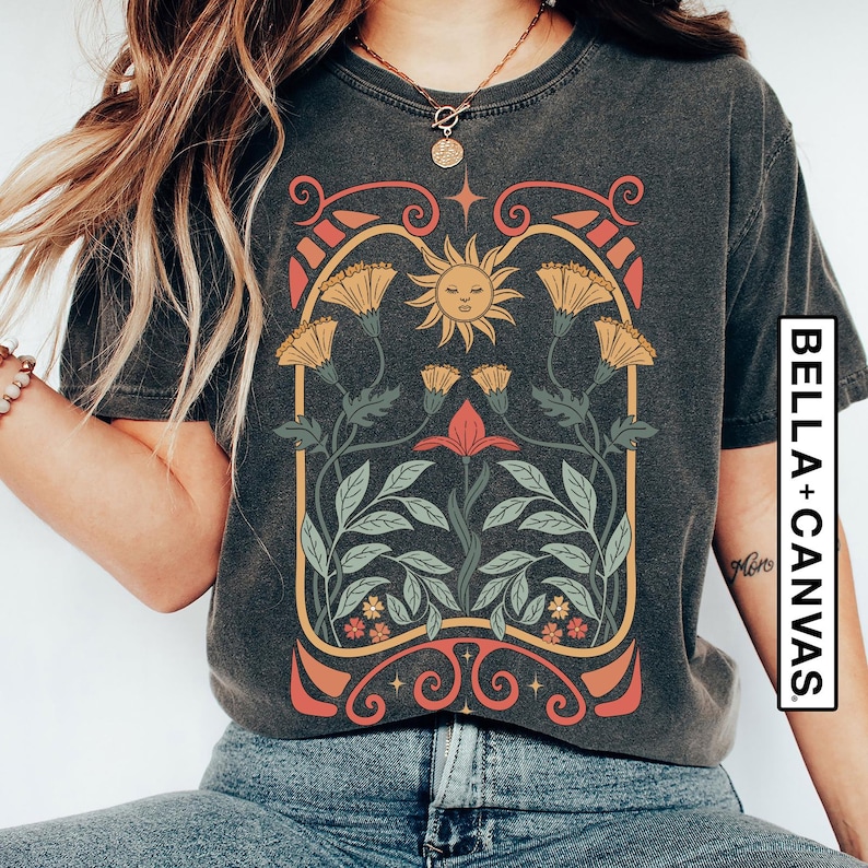 May include: A dark grey t-shirt with a vintage-style graphic design featuring a sun with a face, flowers, and leaves. The design is framed by a decorative border.