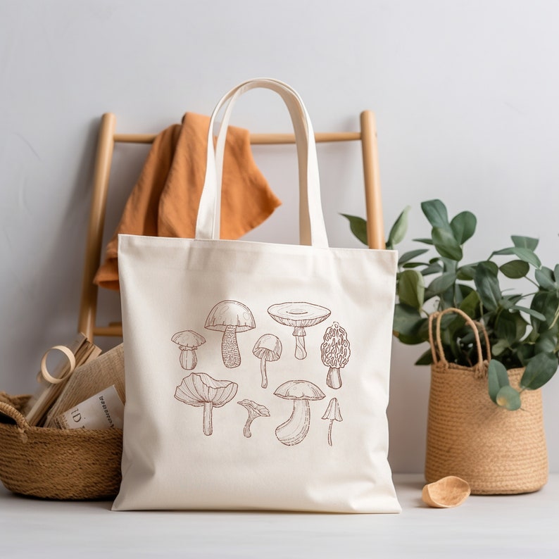 May include: A white canvas tote bag with brown line drawings of various mushrooms. The bag has two handles and is sitting on a wooden surface.