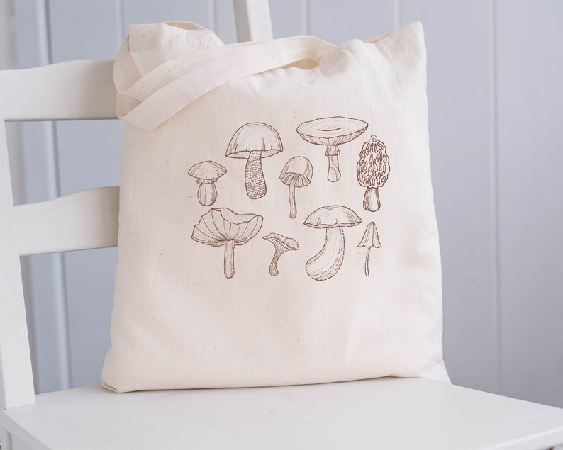 May include: A white canvas tote bag with a brown line drawing of various mushrooms. The mushrooms are drawn in a simple, realistic style.