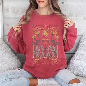 Op de afbeelding: Een rood sweatshirt met een vintage-stijl grafische afbeelding van een zon en bloemen. De afbeelding is in tinten geel, oranje en groen.