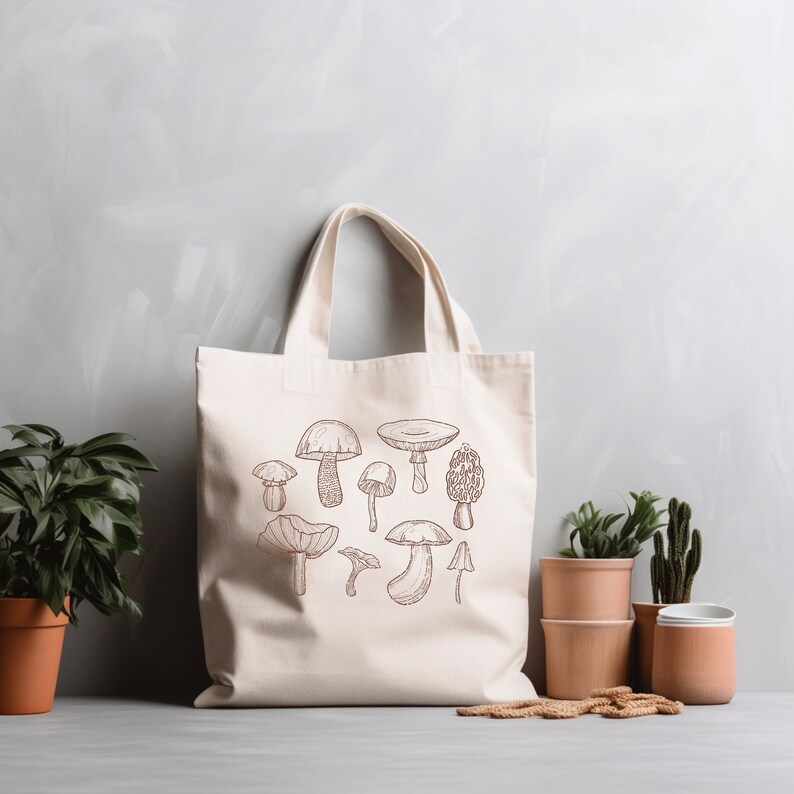 May include: A white canvas tote bag with a brown line drawing of various mushrooms. The bag has two handles and is sitting on a gray surface.