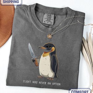 May include: A charcoal grey t-shirt featuring a graphic of a penguin holding a knife with the text FLIGHT WAS NEVER AN OPTION printed below. The shirt has a crew neckline and a Comfort Colors brand label on the inside of the collar.