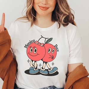 May include: White t-shirt with a graphic of two red cherries with faces. The cherries are holding hands and have the names "Monica" and "Dax" written on them.