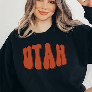 May include: Black sweatshirt with the word "UTAH" in a retro, orange font. The sweatshirt is a crew neck style. The image shows a person wearing the sweatshirt.