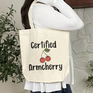 Peut inclure: Un sac fourre-tout en toile blanche avec le texte "Certified Armcherry" et un dessin de deux cerises rouges avec des tiges et des feuilles vertes.