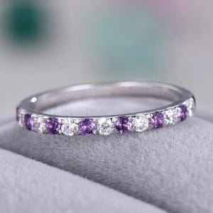 Peut inclure: Une bague en argent ornée de pierres précieuses violettes et transparentes alternées. La bague présente une rangée de petites pierres précieuses rondes violettes entrecoupées de pierres claires et scintillantes. La bague est posée sur une surface grise.