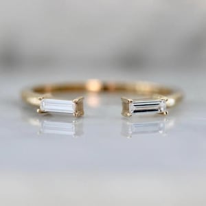 Op de afbeelding: Een gouden ring met twee baguette-geslepen diamanten die in de band zijn gezet. De ring is aan één kant open, waardoor het een uniek en modern ontwerp is.