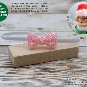 May include: A baby pink headband with a sparkly bow, displayed on a wooden block. The headband is white and the bow is covered in small, shiny embellishments. A green circle with the text "BABY PINK HEADBAND" is in the upper left corner.