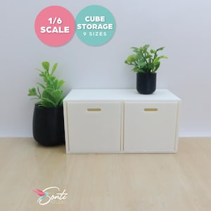 May include: A miniature white cube storage unit with two drawers, each featuring a small rectangular handle. Two small potted plants with green foliage sit on and beside the unit. The text reads "1/6 SCALE" and "CUBE STORAGE 9 SIZES".
