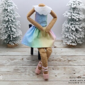 May include: A doll wearing a rainbow-coloured dress with a white collar and pink ballet shoes. The doll is sitting on a small black chair. Two miniature snow-covered Christmas trees are in the background.