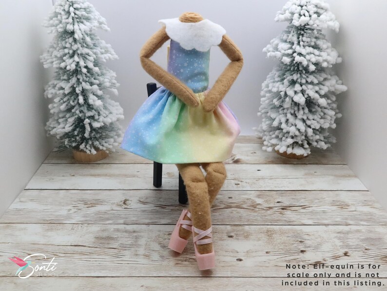 May include: A doll wearing a rainbow-coloured dress with a white collar and pink ballet shoes. The doll is sitting on a small black chair. Two miniature snow-covered Christmas trees are in the background.