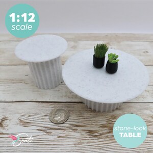 May include: Miniature white stone-look table and side table, 1:12 scale. The round table has two small black pots with green plants. The side table is a smaller version of the table. A dime is next to the tables.