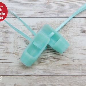 May include: A pair of mint green sparkle shoes with light blue ribbons, set against a light wood background. The shoes are small, possibly for a child or decorative use. A red circle with white text reads "Mint Sparkle Shoes".