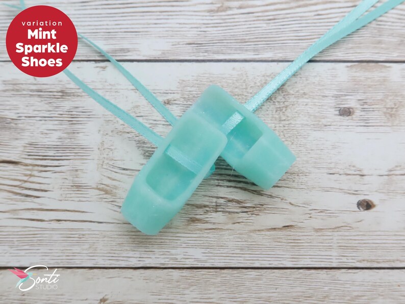 May include: A pair of mint green sparkle shoes with light blue ribbons, set against a light wood background. The shoes are small, possibly for a child or decorative use. A red circle with white text reads "Mint Sparkle Shoes".