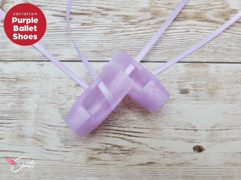 May include: Two lilac ballet shoes with ribbons, set on a light wood surface. The shoes have white straps and a satin finish. The text "Purple Ballet Shoes" is in a red circle.