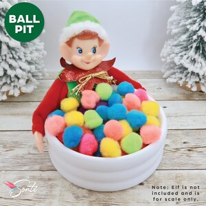 May include: A white plastic ball pit filled with colorful pom-poms. A small elf doll is sitting in the ball pit. The text "BALL PIT" is written in black on a green circle.