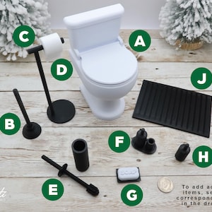 May include: A miniature white toilet with a closed lid, surrounded by black bathroom accessories. Includes a toilet paper holder, plunger, soap, and a black bath mat. The text in the image says "To add additional items, select the corresponding letter in the drop down."