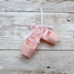 May include: A pair of pale pink ballet slippers with matching ribbons tied in bows. The slippers are made of a solid material and are designed to resemble traditional ballet shoes. The background is a light wood grain.