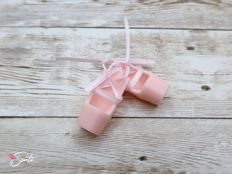 May include: A pair of pale pink ballet slippers with matching ribbons tied in bows. The slippers are made of a solid material and are designed to resemble traditional ballet shoes. The background is a light wood grain.