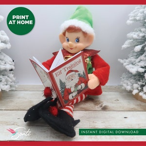 Könnte beinhalten: Eine Elfenpuppe mit grünem Hut sitzt auf einer Holzoberfläche und liest ein Buch mit dem Titel "Elf Training". Das Buch hat einen rot-weiß gestreiften Einband mit einer Illustration des Weihnachtsmanns.