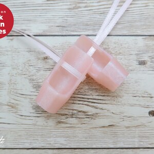 May include: Pale pink satin ballet shoes with white ribbons, set against a light wood grain background. The shoes have a strap across the top. The text "Pink Satin Shoes" is in a red circle. The shoes are a delicate, pastel colour.