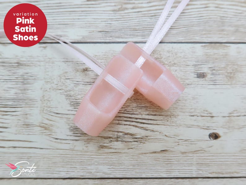 May include: Pale pink satin ballet shoes with white ribbons, set against a light wood grain background. The shoes have a strap across the top. The text "Pink Satin Shoes" is in a red circle. The shoes are a delicate, pastel colour.