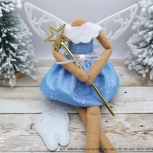 May include: A blue fairy doll with white wings, a gold star wand, and a white tooth charm. The doll is wearing a blue dress with white stars and a white sash. The doll is sitting on a wooden surface with a white tooth prop.