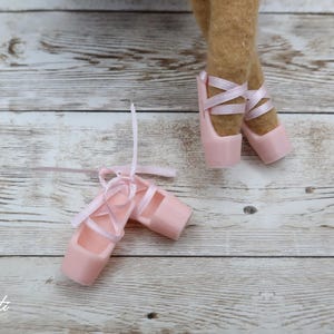 May include: A pair of pink ballet pointe shoes with satin ribbons, next to a pair of legs wearing similar shoes. The shoes are on a light-coloured wooden surface. The shoes are a soft pink colour.