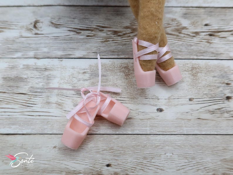 May include: A pair of pink ballet pointe shoes with satin ribbons, next to a pair of legs wearing similar shoes. The shoes are on a light-coloured wooden surface. The shoes are a soft pink colour.