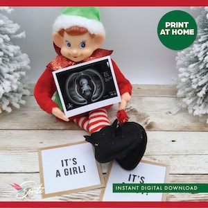 May include: An elf doll holding an ultrasound image, with the text "IT'S A GIRL!" on a letter board. The image also includes a sign that says "PRINT AT HOME" and "INSTANT DIGITAL DOWNLOAD". The elf is wearing a red and green outfit.